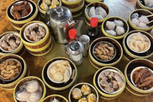 A round table set with various bamboo steamers filled with dim sum dishes, including dumplings, buns, and meats. A teapot, soy sauce bottles, and a metal shaker are in the center.