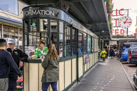 People gather near a market information booth in an outdoor shopping area. A large clock with red neon letters is visible. The setting includes a mix of buildings and parked cars, with flower baskets hanging from the eaves.