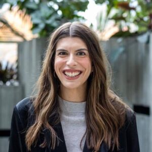 A woman with long brown hair smiles while standing in an outdoor setting with greenery in the background. She is wearing a black blazer over a grey top.