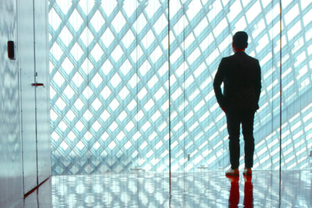 A person stands silhouetted in a corridor with a high ceiling, facing a large grid-patterned window. The polished floor reflects the figure, creating a serene and contemplative atmosphere.