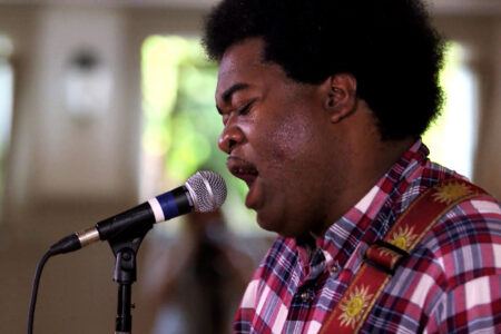 A man with an afro hairstyle sings into a microphone. He is wearing a red checkered shirt with floral designs on the strap of his guitar. The background is blurred, emphasizing his singing expression.