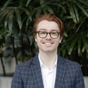 A smiling person with red hair wearing glasses, a white shirt, and a blue plaid blazer stands in front of green leafy plants.