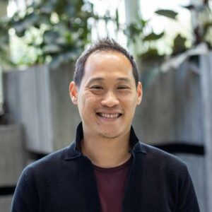 A person is smiling while standing in an indoor setting. They are wearing a black jacket over a maroon shirt. The background features blurred greenery and concrete structures.