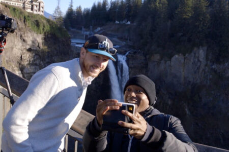 Two people stand on a viewing platform taking a selfie with a smartphone. Behind them, a waterfall cascades down a rocky cliff, surrounded by trees. A tripod with a camera is set up on the railing nearby.