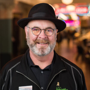 A man with a gray beard and waxed mustache, wearing glasses and a black hat, smiles in an indoor setting with blurred lights in the background. He is wearing a black jacket with a name tag.