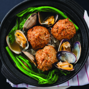 A clay pot dish featuring meatballs, clams, and glass noodles, garnished with green vegetables. The dish is served on a black surface with a striped cloth underneath.