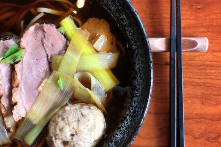 A bowl of noodle soup with slices of meat, vegetables, and a meatball, served in a black bowl on a wooden table. Black chopsticks are placed on the right side of the bowl.