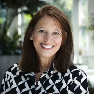 A woman with shoulder-length brown hair smiles warmly at the camera. She is wearing a black and white patterned blouse. The background features soft-focus greenery indoors, suggesting a modern, bright environment.