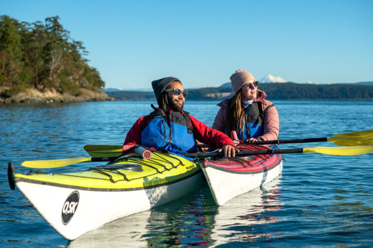 Two people kayaking on a calm, clear blue lake under a sunny sky. Both wear beanies, sunglasses, and life vests. Forested shoreline and distant mountains are visible in the background.