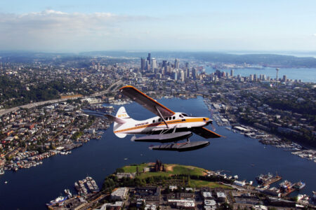 A seaplane flies over the bustling city of Seattle, with its soaring skyscrapers and expansive lake below. The skyline is dotted with tall buildings while green spaces and winding roads lay beneath a clear sky, offering a bird's-eye view of exciting things to do in this vibrant metropolis.