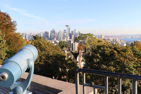 A scenic view of a cityscape with the Seattle Space Needle in the distance. Trees and buildings are in the foreground, and a blue telescope is positioned on the left side, overlooking the city under a clear blue sky.