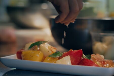 A hand sprinkles salt over a salad composed of colorful tomatoes, white cheese cubes, basil leaves, and thin slices of onion on a white rectangular plate. A blurred background with a pot suggests a kitchen setting.