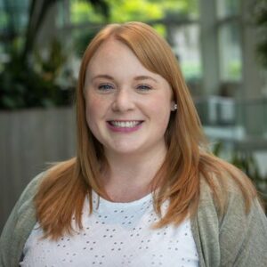 A woman with long red hair smiles warmly at the camera. She is wearing a white eyelet blouse and a gray cardigan. The background is a bright, modern interior with large windows and greenery.