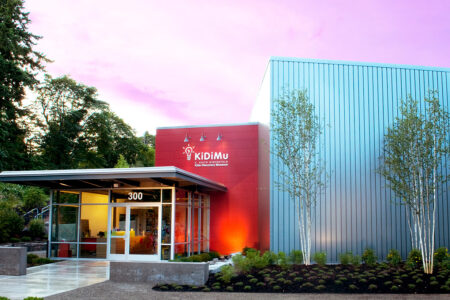 A modern building with a red façade and silver siding, featuring the Kidimu logo and slogan. It is surrounded by trees and landscaping, with a purple-pink sky at sunset in the background.