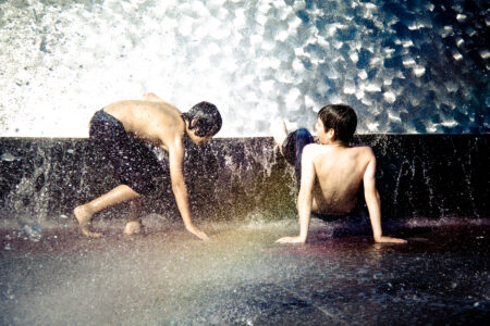 Two children play joyfully in a cascading water fountain. One crouches, the other sits on the wet surface, both enjoying the splash and sunlight. The water glimmers in the background, creating a lively and refreshing scene.
