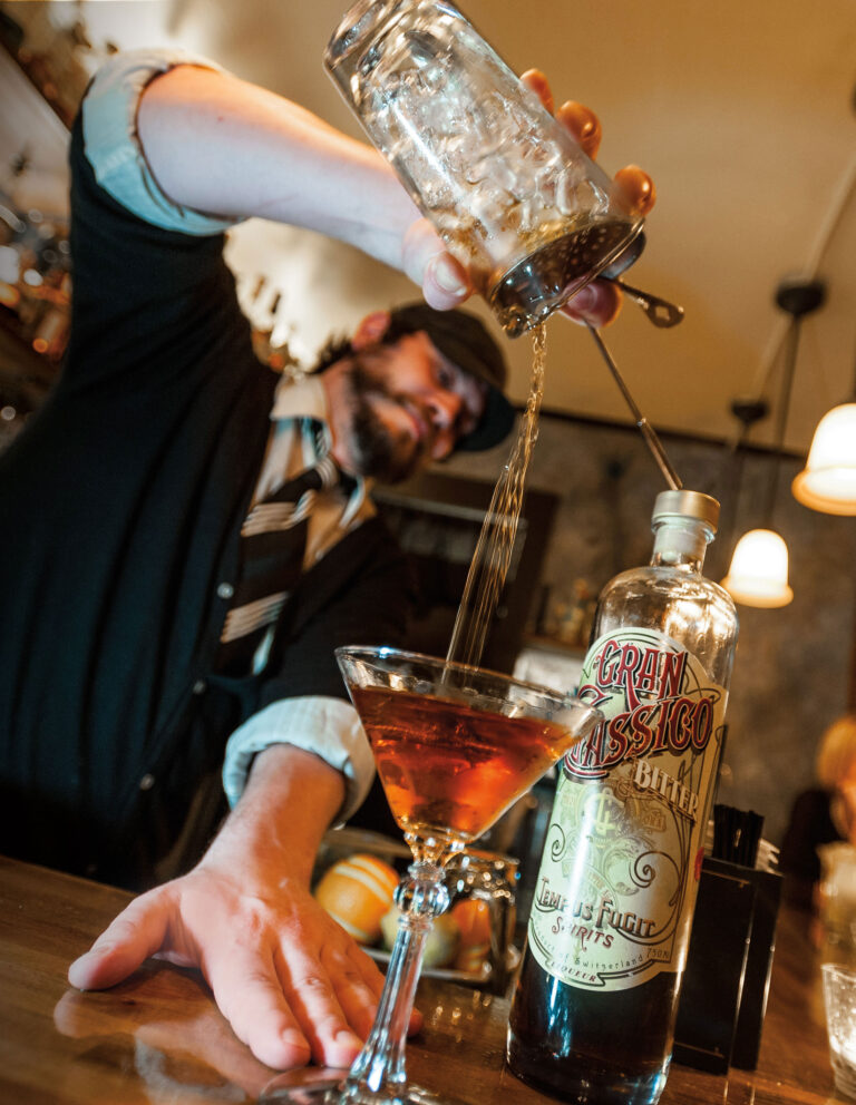 A bartender expertly pouring a drink from a cocktail shaker into a martini glass on a bar. A bottle labeled "Gran Classico" is in the foreground. The bartender is wearing a hat and tie, and the bar is warmly lit.