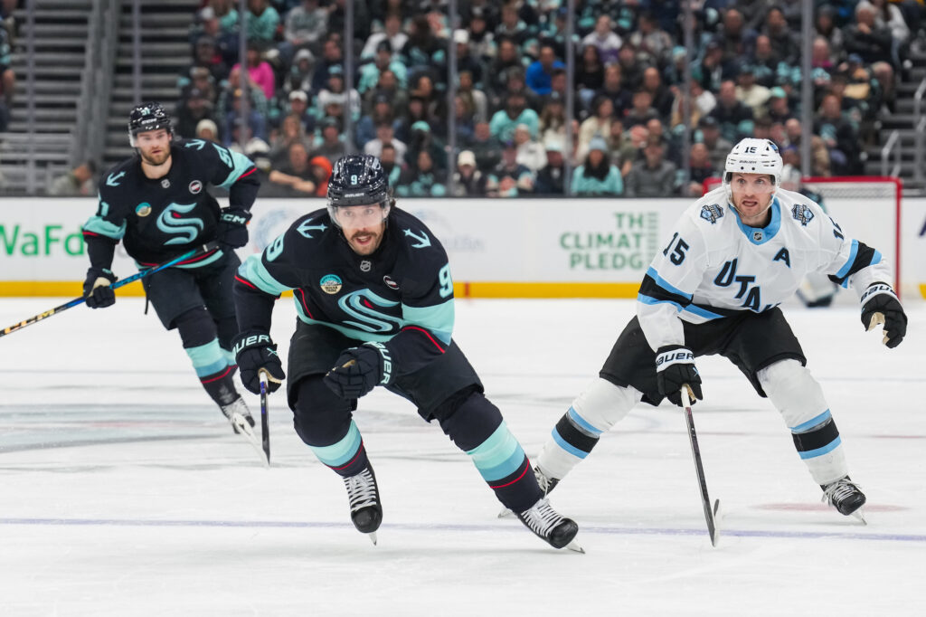 Hockey players in action on the ice rink evoke the dynamic spirit of Seattle sports. Two players in dark jerseys with teal accents skate toward the puck, while a player in a white and blue jersey follows behind. The audience is visible in the background.