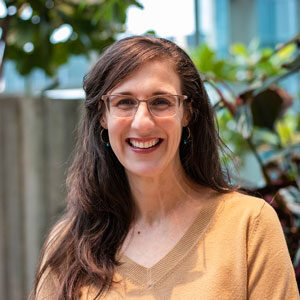 A person with long brown hair smiles while wearing glasses and a beige sweater. They are standing in front of a blurred background featuring green plants.