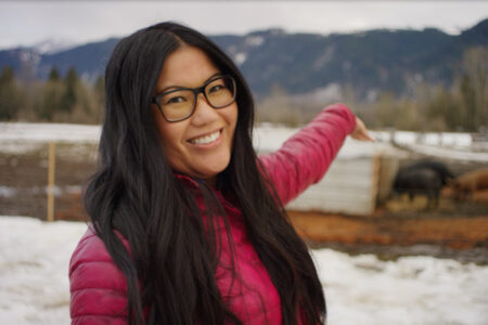 A smiling person with long black hair and glasses wearing a pink jacket points towards a snowy field with mountains in the background. There are farm animals and a small shed visible in the distance.