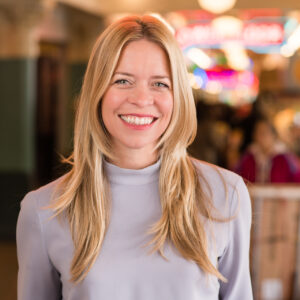 Smiling woman with long blonde hair wearing a light gray blouse stands indoors. The background is softly blurred, featuring colorful lights and people in a bustling environment.