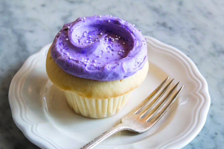 A vanilla cupcake with swirled purple frosting and white sprinkles sits on a white plate. A silver fork rests beside the cupcake on a marble surface.