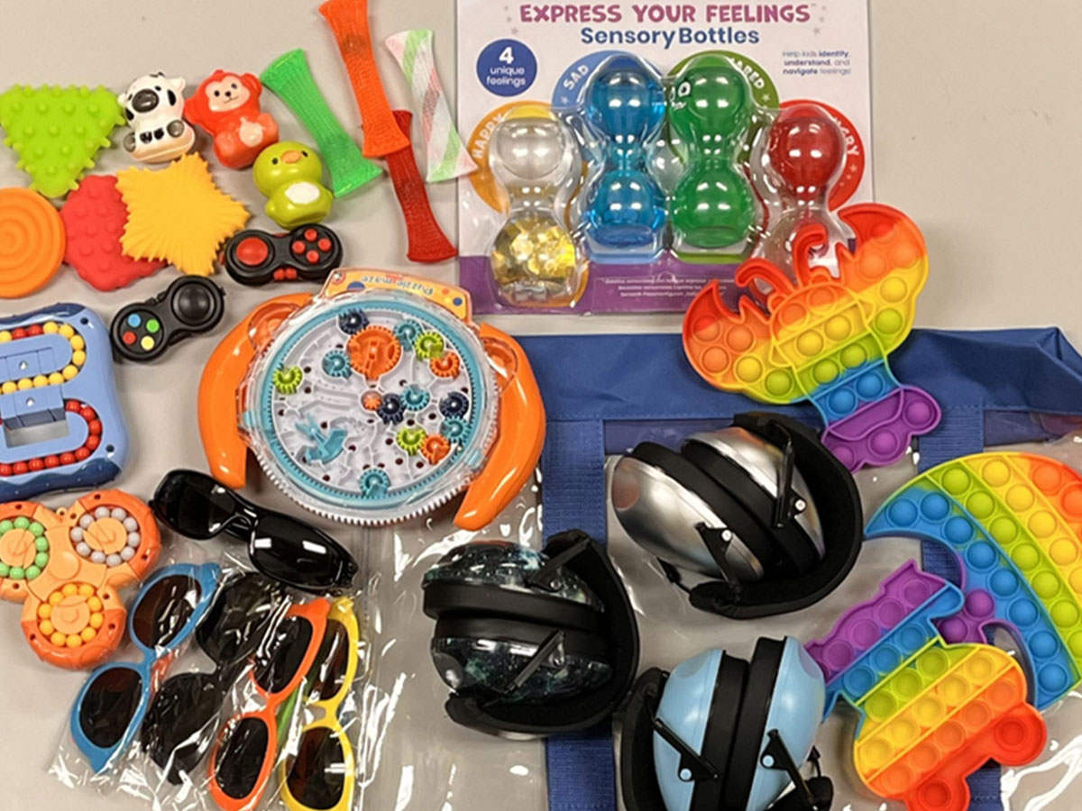 A variety of sensory toys and tools are spread out on a surface. Items include colorful textured toys, pop-it toys, sensory bottles, spinning toys, sunglasses with different colored frames, and noise-reducing headphones.