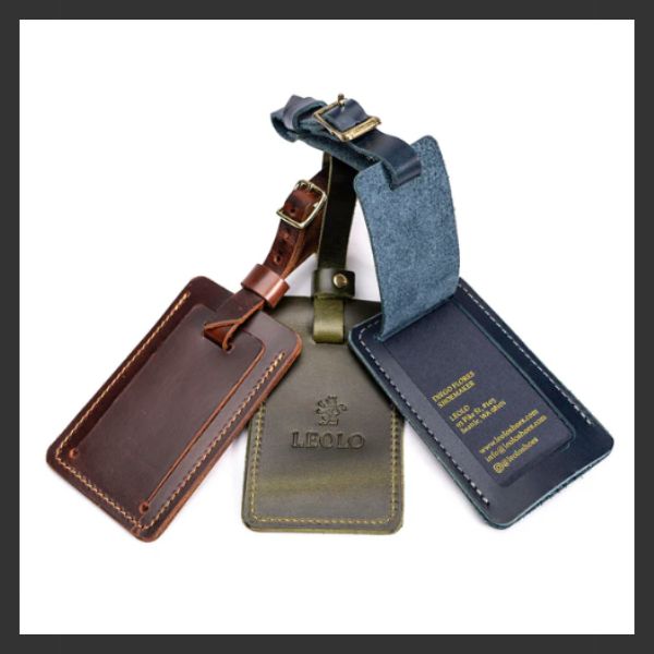 Three leather luggage tags in brown, green, and navy blue, each with visible stitching and buckled straps, are arranged overlapping on a white background. One tag shows embossed contact information in gold.