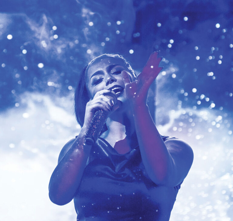 A singer passionately performs onstage, holding a microphone. The background is filled with ethereal, star-like patterns and a blue tint envelops the scene, creating a dreamy atmosphere.