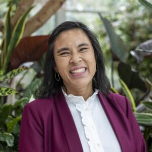 A person wearing a maroon blazer and white blouse stands smiling in front of a background filled with lush green plants.