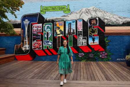 A woman in a teal dress stands on a wood platform in front of a mural painted on the wall behind her that reads 