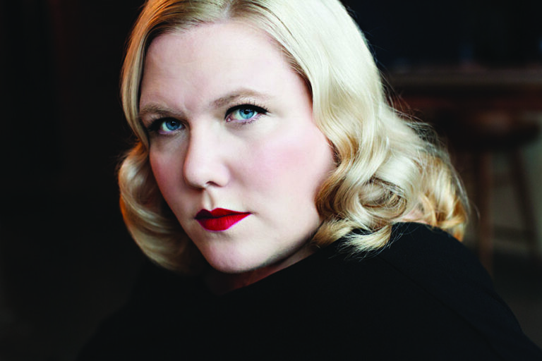 A person with wavy, blonde hair and bright red lipstick gazes seriously at the camera. The background is blurred, creating a moody and focused portrait effect.