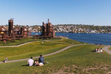groups of people lounging in the grass with the steampunk-esque towers of Gas Works Park rise in the distance, the waters of Lake Union beyond.