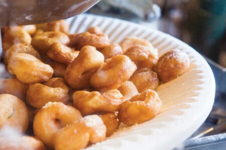 A close-up of a plate filled with small, golden-brown donuts. The donuts have a shiny, glazed surface and are piled on a white paper plate. The image captures the warm texture and appealing appearance of the freshly made treats.