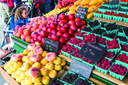 A bustling farmers market with fresh produce on display, including peaches, nectarines, raspberries, and boysenberries. Shoppers browse the stands, with a mix of adults and children visible in the busy scene.
