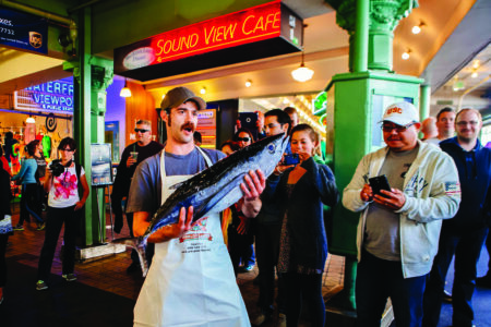 A man wearing an apron holds a large fish in a bustling indoor market. Onlookers gather around, some taking pictures. The backdrop features green pillars and a neon sign that reads 