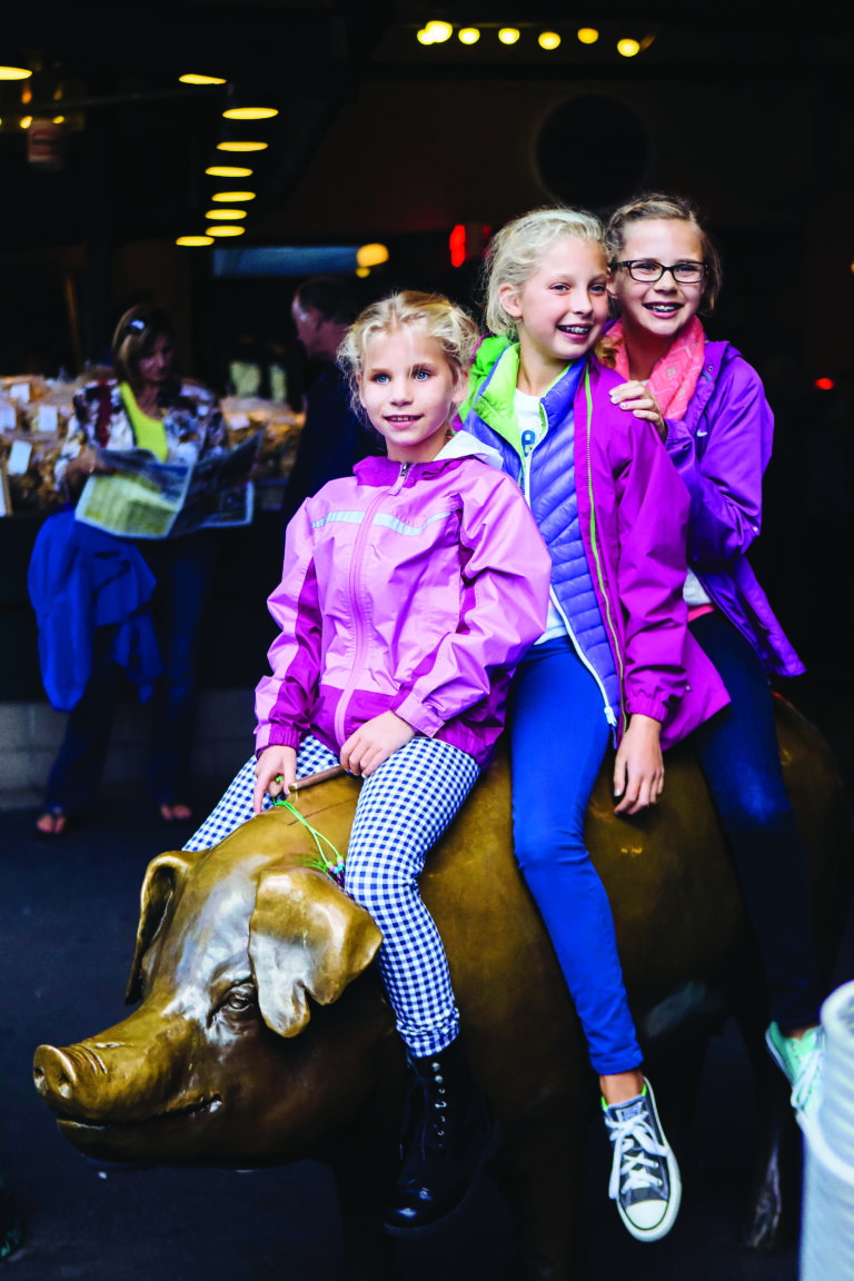 Three children dressed in colorful jackets are joyfully sitting on a bronze pig statue. The background shows a market setting with people and shelves. The children are smiling and appear to be enjoying their time.