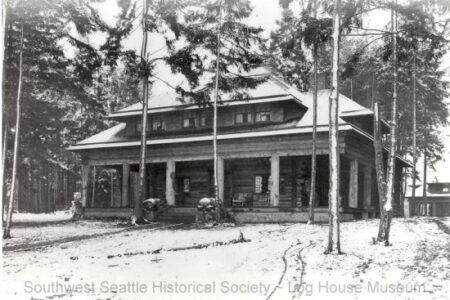 A historical black-and-white photo of a large log house surrounded by tall trees and snow on the ground. The house has a covered front porch and multiple windows. It is part of the Southwest Seattle Historical Society.