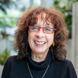 Smiling woman with curly brown hair wearing red glasses and a black turtleneck, standing indoors with blurred greenery in the background.