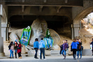 A large troll statue under a concrete bridge is draped with colorful sports jerseys. Several people stand around, some posing for photos and others walking or talking nearby.