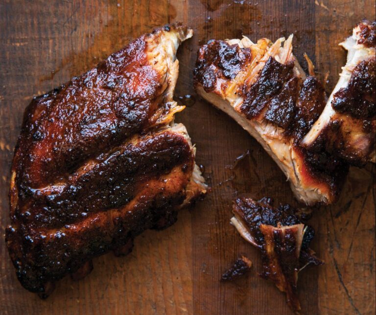 Delicious barbecued ribs with a dark, caramelized crust are laid out on a wooden cutting board. Some of the meat is pulled apart, revealing juicy, tender insides.