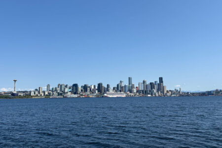 Seattle skyline view from the water on a clear day, featuring the Space Needle on the left, tall downtown buildings, and a large white cruise ship docked near the shore, with blue sky and sea in the foreground.