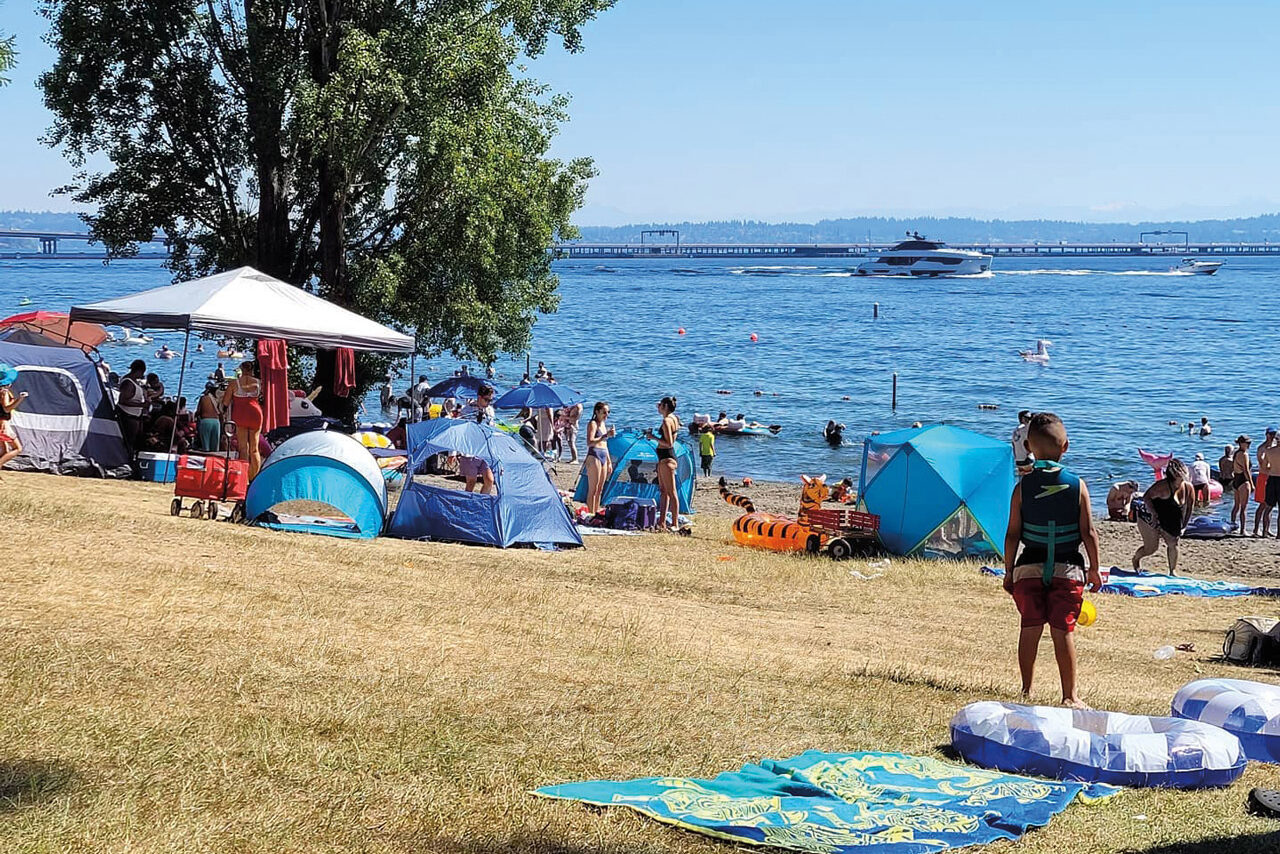 a boy in a life vest stands on a grassy beach with scattered beach umbrellas, gazing toward the water