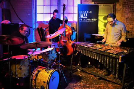 Three musicians perform in a cozy venue with a brick wall backdrop. A drummer, a double bassist, and a vibraphonist play together. A 