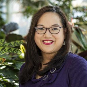 A person with long dark hair and glasses smiles while wearing a purple sweater. They stand in front of lush green plants, with natural light streaming in from a window.