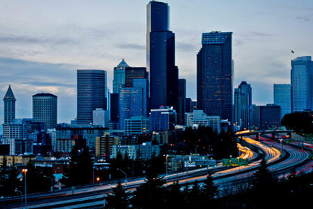 City skyline at dusk with tall skyscrapers and a busy highway in the foreground, displaying streaks of car lights. The sky is overcast, adding a muted ambiance to the urban scene.