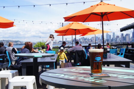 An outdoor patio with orange umbrellas and tables, overlooking a city skyline. People are seated and standing, enjoying the view. A glass of beer is prominently displayed on a table in the foreground. String lights are hanging overhead.