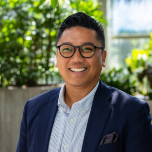 A person in glasses and a navy blazer smiles warmly. They stand against a backdrop of lush green plants in a bright, sunlit space.