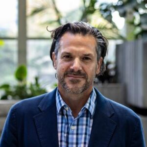 A person with a beard and wavy hair wearing a blue blazer over a checkered shirt stands indoors. The background includes large windows and green plants, creating a natural, relaxed atmosphere.