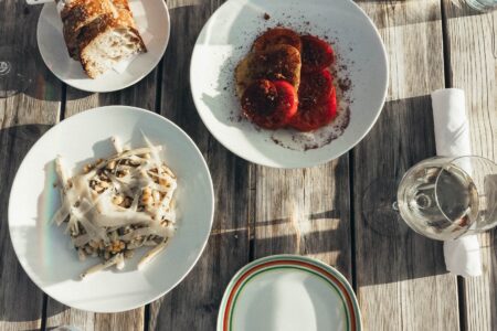 A wooden table set with plates of sliced bread, a pasta dish with shaved cheese and pine nuts, a plate of sliced tomatoes and seasoned fried rounds, a wine glass, and a rolled napkin.