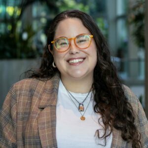 A person with long, curly hair and orange glasses is smiling. They are wearing a plaid jacket over a white shirt, accessorized with necklaces. The background is blurred, with hints of plants and glass.